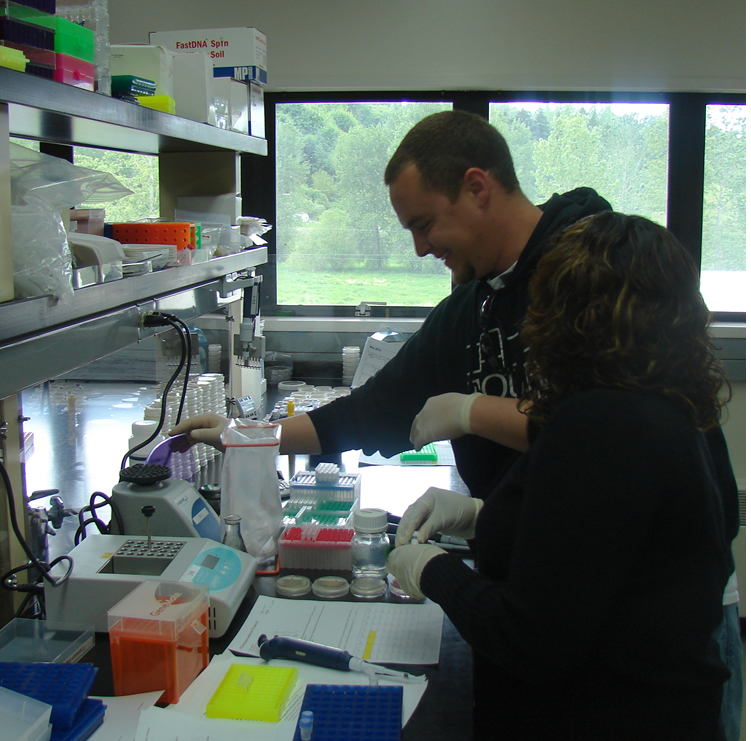 Two students at a lab bench. The student further from the camera is closing the purple lid of a mini centrifuge, the rest of which is hidden behind a vortexer. On the bench in front of the student closer to the camera are some small petri dishes, a paper with highlighted text, a pipette laying on its side, and a "dry bath" for heating microcentrifuge tubes to specified temperatures. On the bench to the right of the vortexer is an orange frame with an autoclavable bag attached at the top.