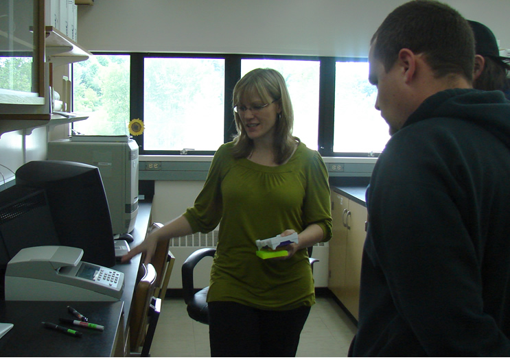 a woman in a green shirt holds two trays for small tubes in one hand as she gestures at a machine out of sight on the counter in front, demonstrating to the student looking in from far right of the frame how to set the thermocycler.