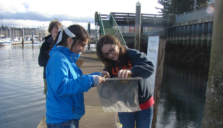 Two students holding a single mesh bait bag with a dowel at the top, both looking at the bag as another person stands behind them.