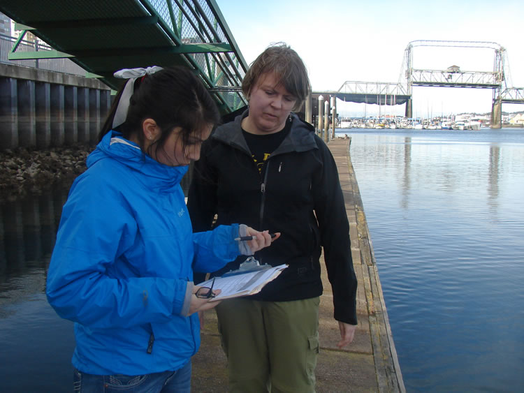 two people standing next to each other, on a dock with a walkway to another level behind them. both are looking at a clipboard held by the person closer to the camera.
