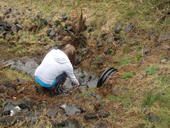 a student in a white hoodie squats by a small black culvert, touching the bit of water that is coming out of it.