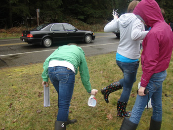 Three students standing together, all of whom are looking away from the camera. The one on the right is bent over a bit, spraying 409 on the soles of the boots of the student in the middle, who has their leg lifted to give access to the spray. The student on the right is facing the first student.
