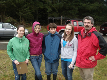 four students and a teacher standing in a row posing for a picture. The student on the left is holding a clipboard facing toward her down by her legs.