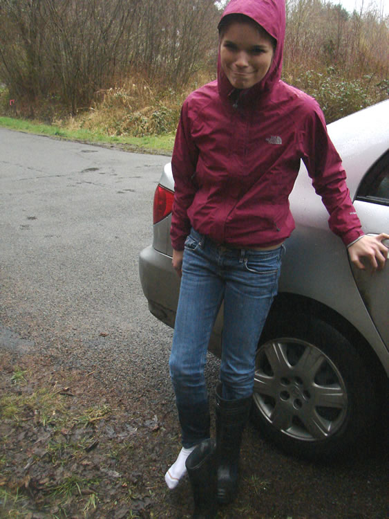 A girl in a pink jacket makes a cheerfully rueful face, standing on her left foot. Her right foot wears a wet white sock, its rubber boot next to it and her jeans wet to the knee