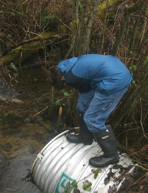 A boy in a blue jacket and black rubber boots stands on a metal culvert.