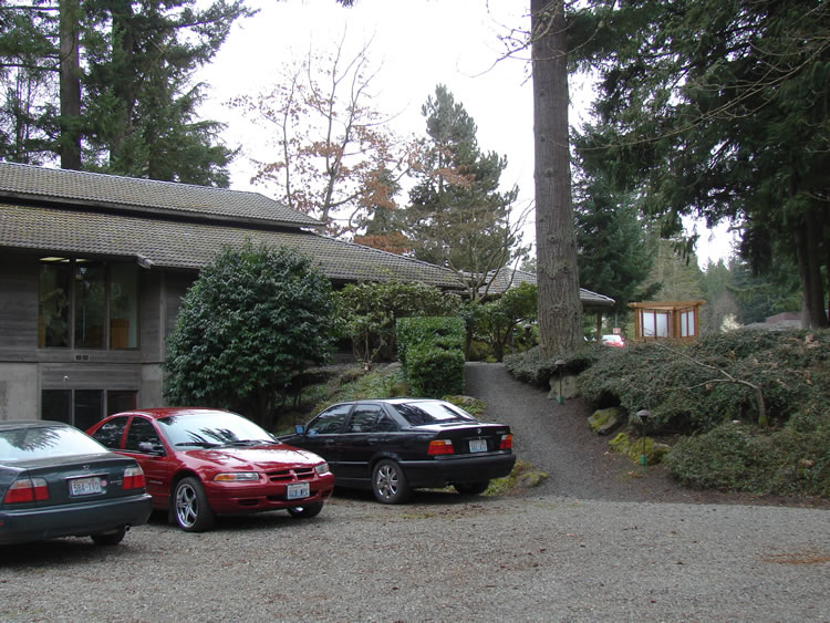 Three cars in a gravel lot outside a two-story gray wooden building. A gravel path leads out of the lot up a small hill.