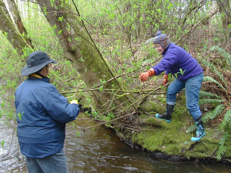 On the left, a woman in a blue jacket and black bucket hat stands in the water of a creek. On the right, a woman in a purple jacket and orange gloves reaches towards the other woman.
