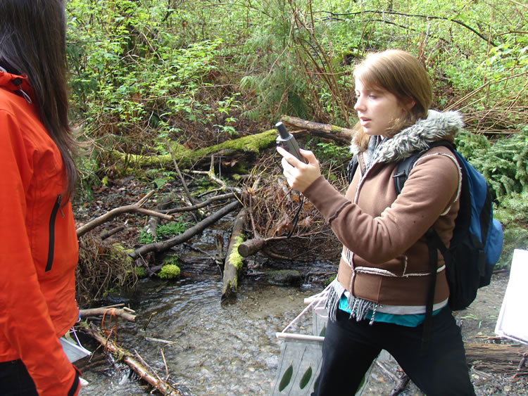 A girl holding a bait bag with bait leaves in it walks through a forest, holding a GPS unit up and looking at the screen.