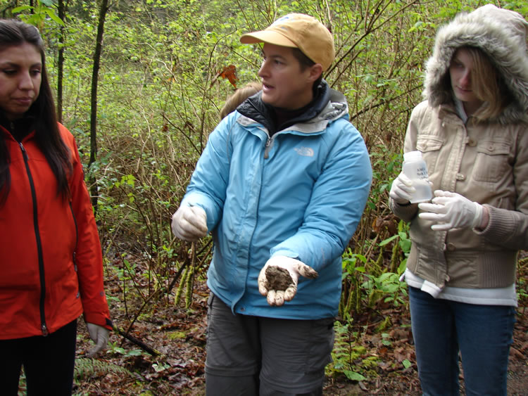 three students standing next to each other. The two girls on the right are wearing latex gloves. The girl in the center holds a ball of soil.
