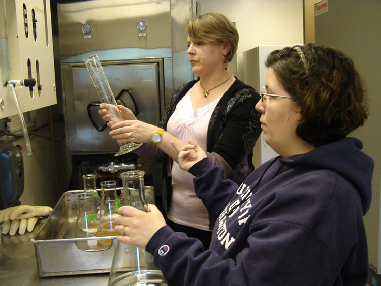 Two women standing next to each other by a metal countertop. The one closer to the camera is holding an Erlenmeyer flask with water in it. The other woman is holding a graduated cylinder. On the counter in front of the second woman is a number of smaller Erlenmeyer flasks, some of which contain an amber liquid.