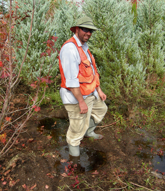 a man in a safety vest and rubber boots demonstrating how deep the water in an unexpected hole in the landscape can be