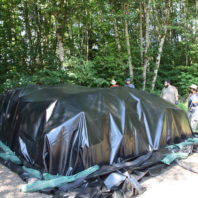 four people standing behind a pile of used pots covered in a large black tarp held down with long green sandbags.