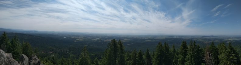 panorama of Puyallup's agricultural area from above and far away. Trees and rocks are in front of the photographer and the sky is blue with streaky white clouds.