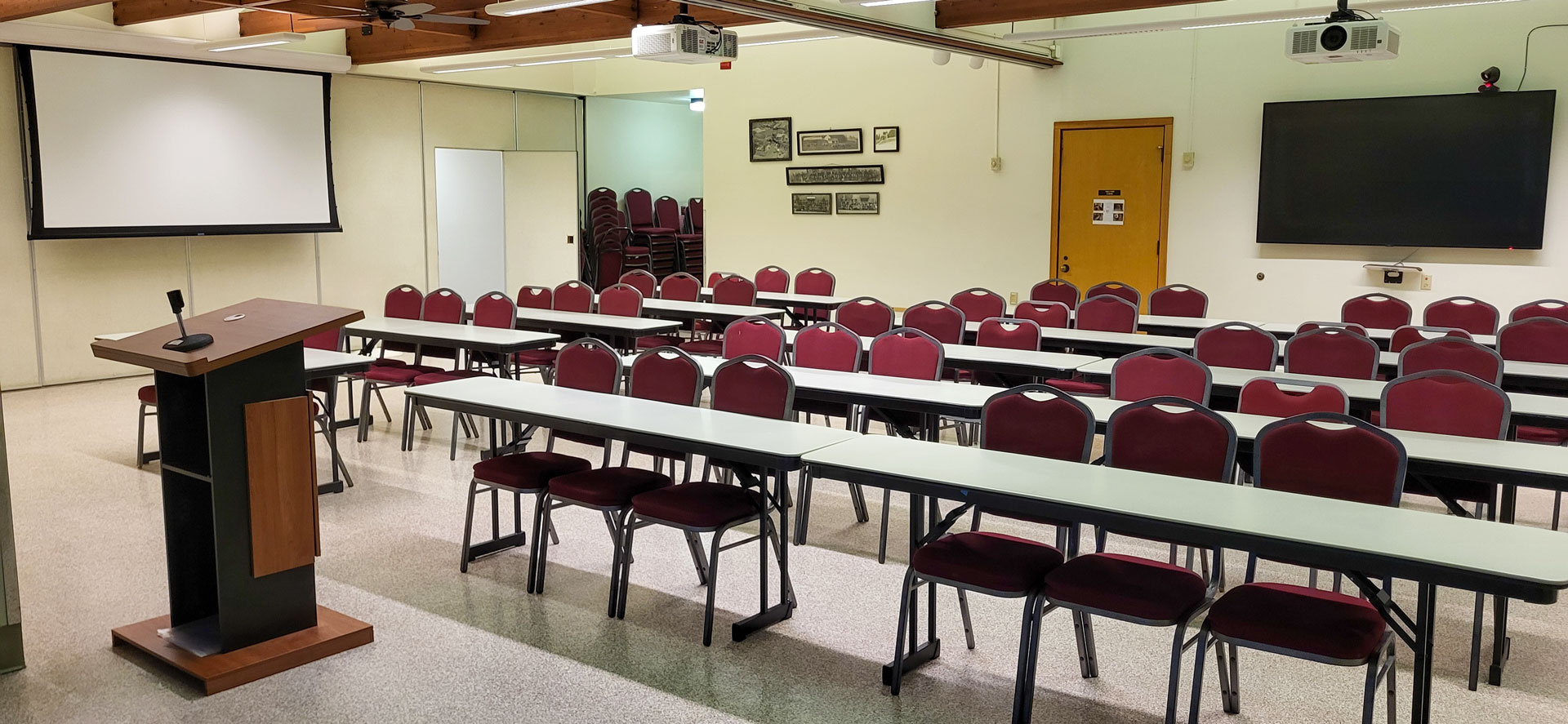 A podium stands in front of tables and chairs in a large meeting room. Projection screen and large monitor on the side and rear walls, respectively.