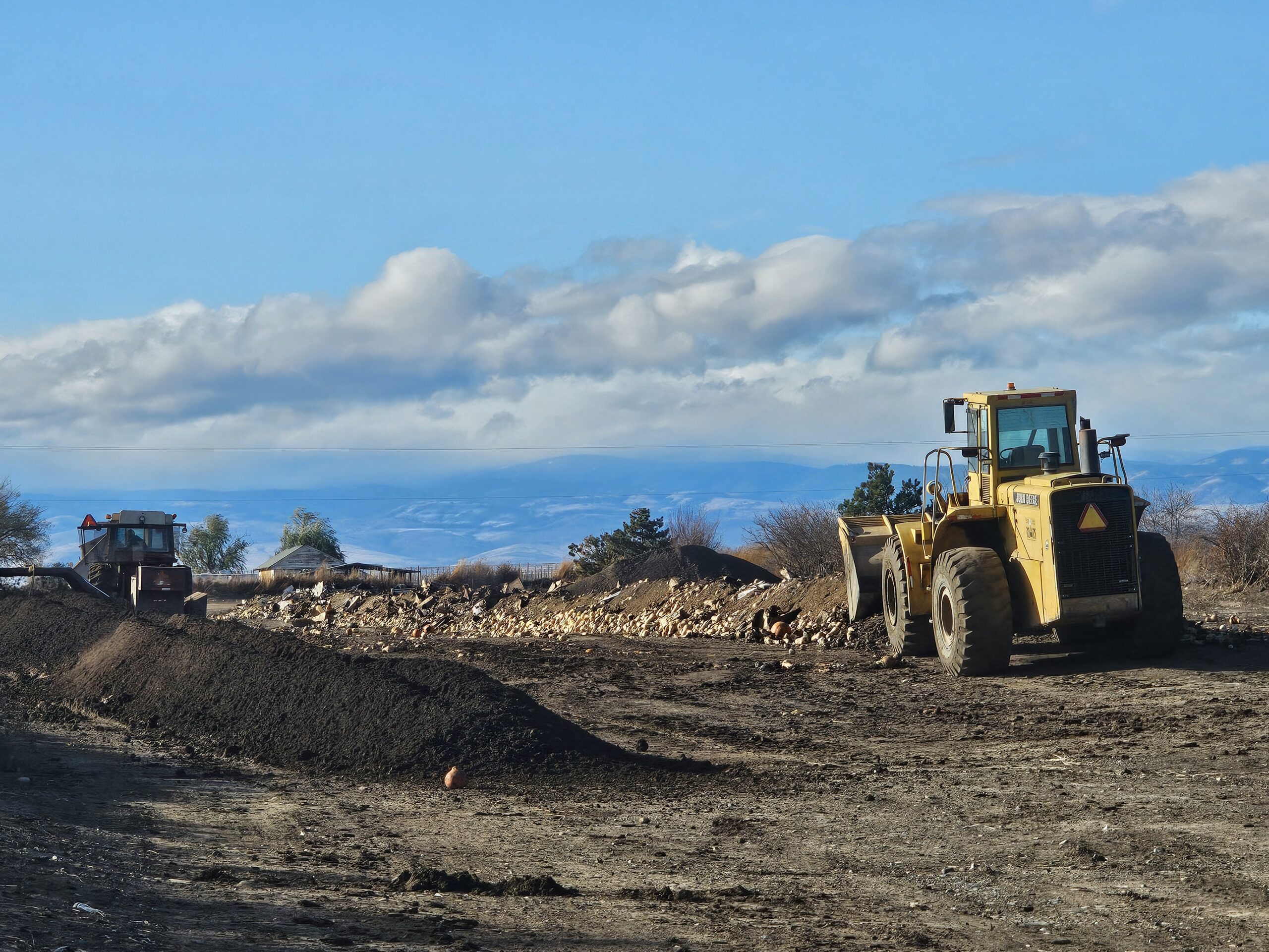 A front loader in an outdoor compost yard with a windrow of finished compost and a window of newly added crop residue and other material.