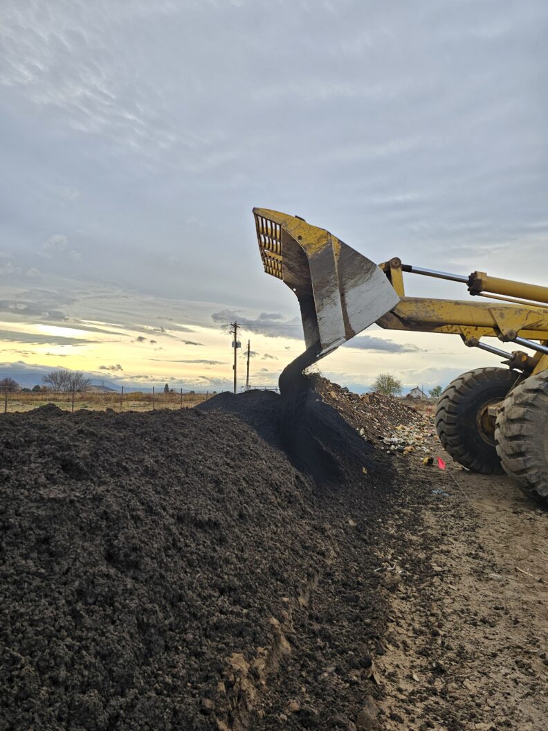 A front loader bucket turning a large compost windrow that is in-process with partially decomposed crop waste and other materials.