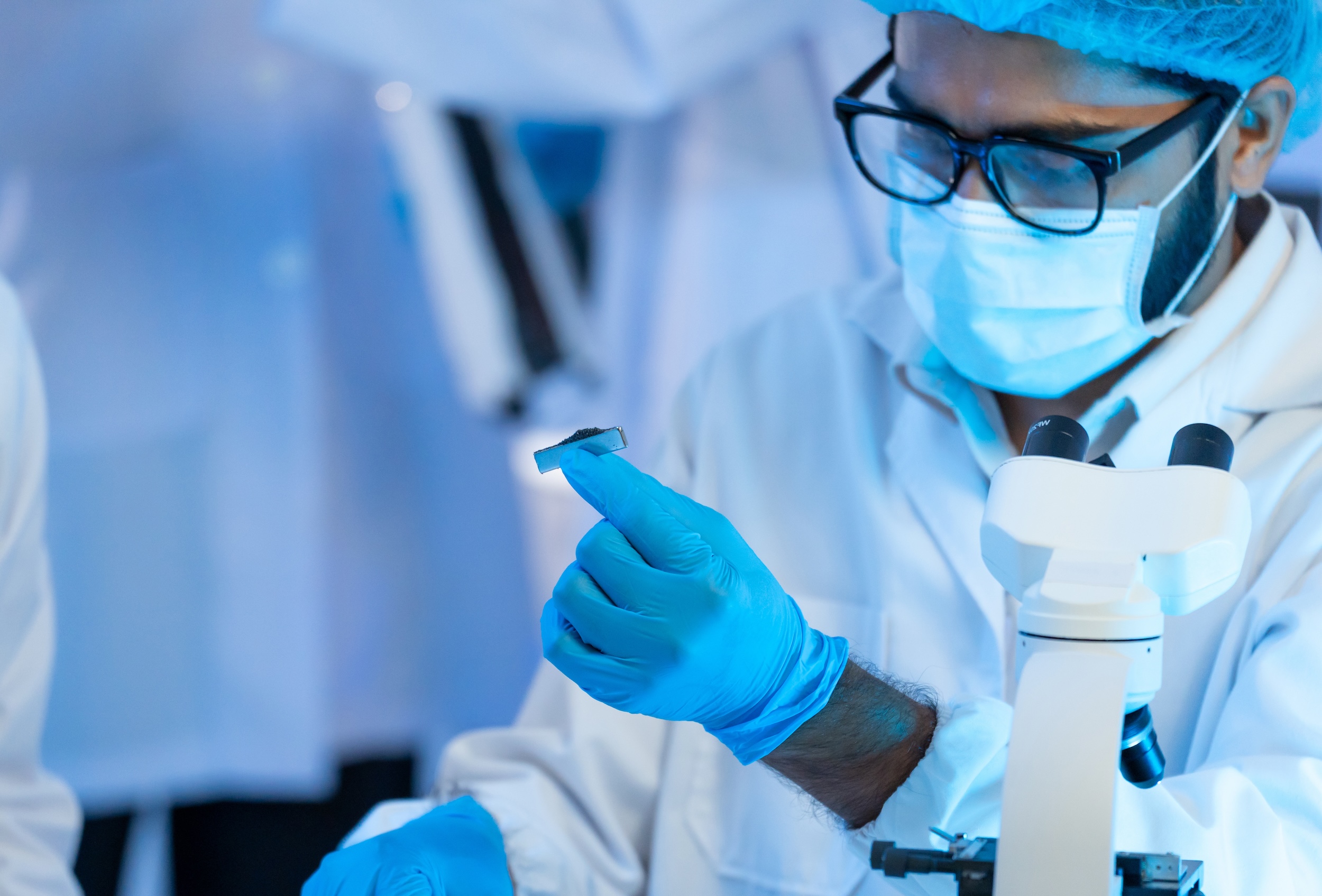Male researcher in mask, glasses and blue gloves examining small specimen