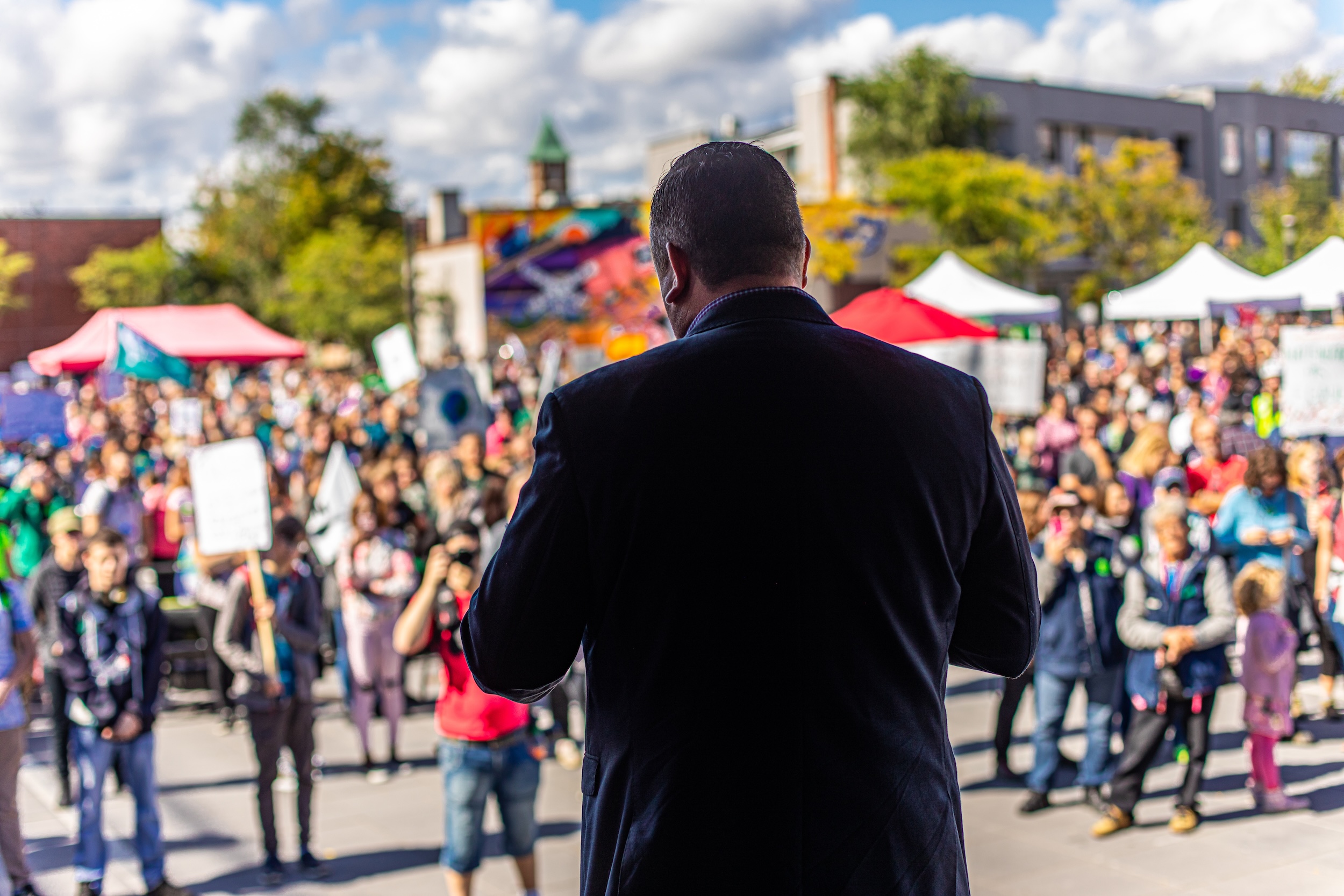 Man on stage from the back taking to crowd at open public space with banners.