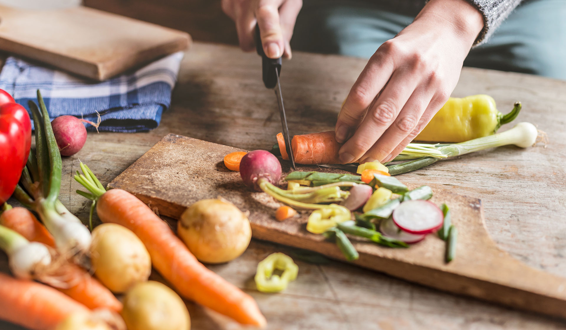 Someone slicing a carrot on a cutting board with other vegetables around the table.