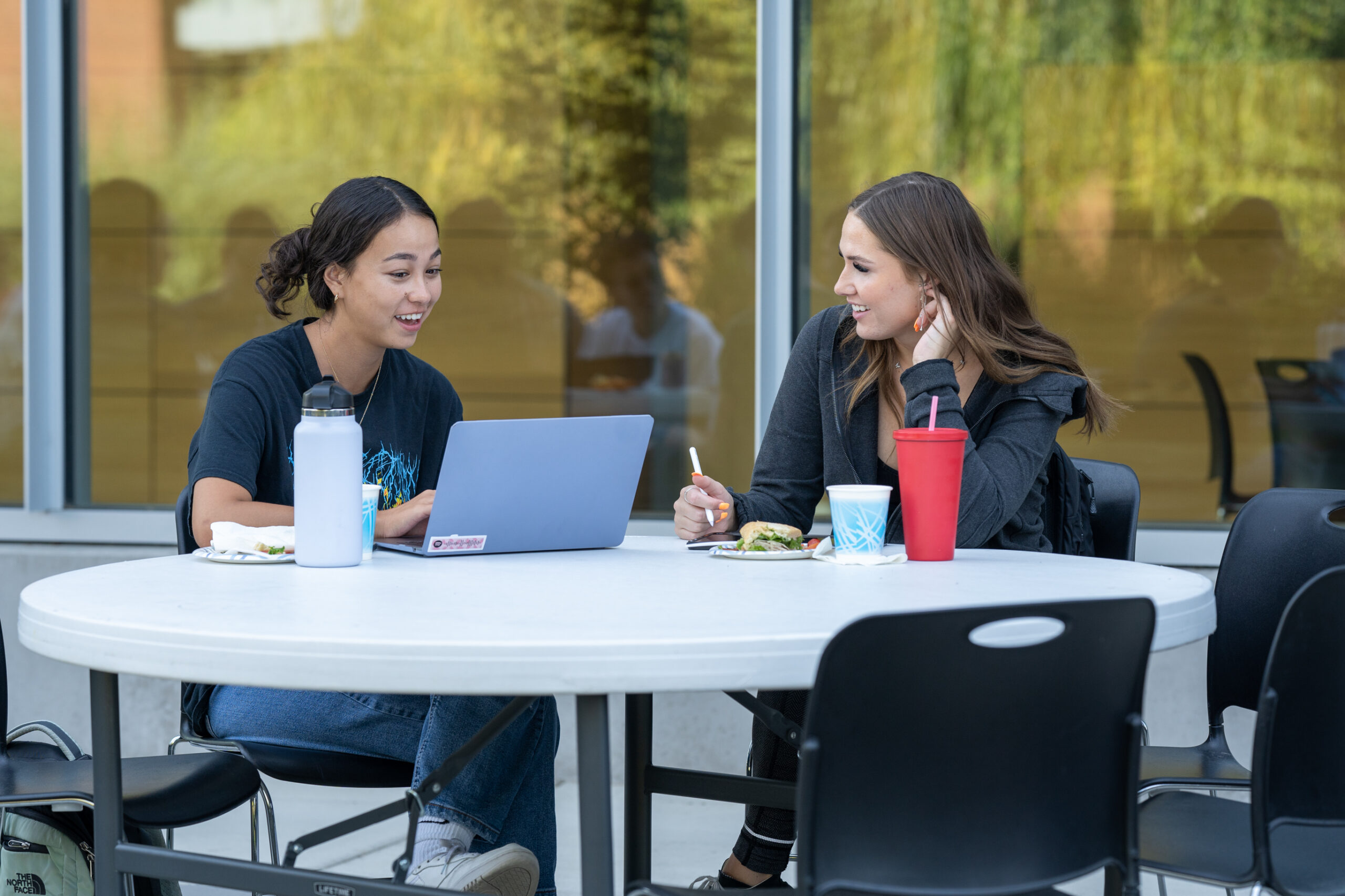 Two students sit outside at a table while working on a computer and writing.