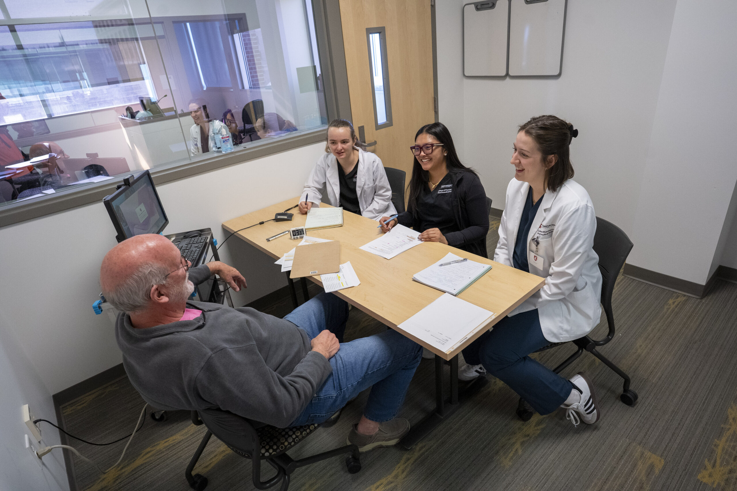 Pharmacy students in white coats doing a mock consultation with a male actor patient.