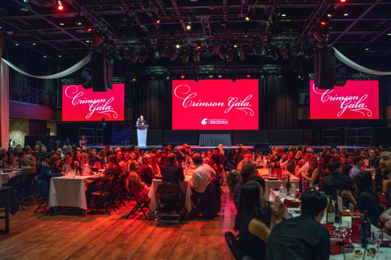 A room filled with people seated at table looking on a stage with Crimson Gala displayed on screens above