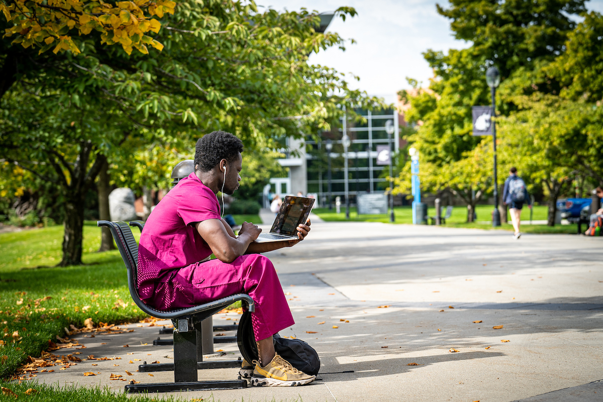 A student sits in their scrubs outside while working on a laptop.