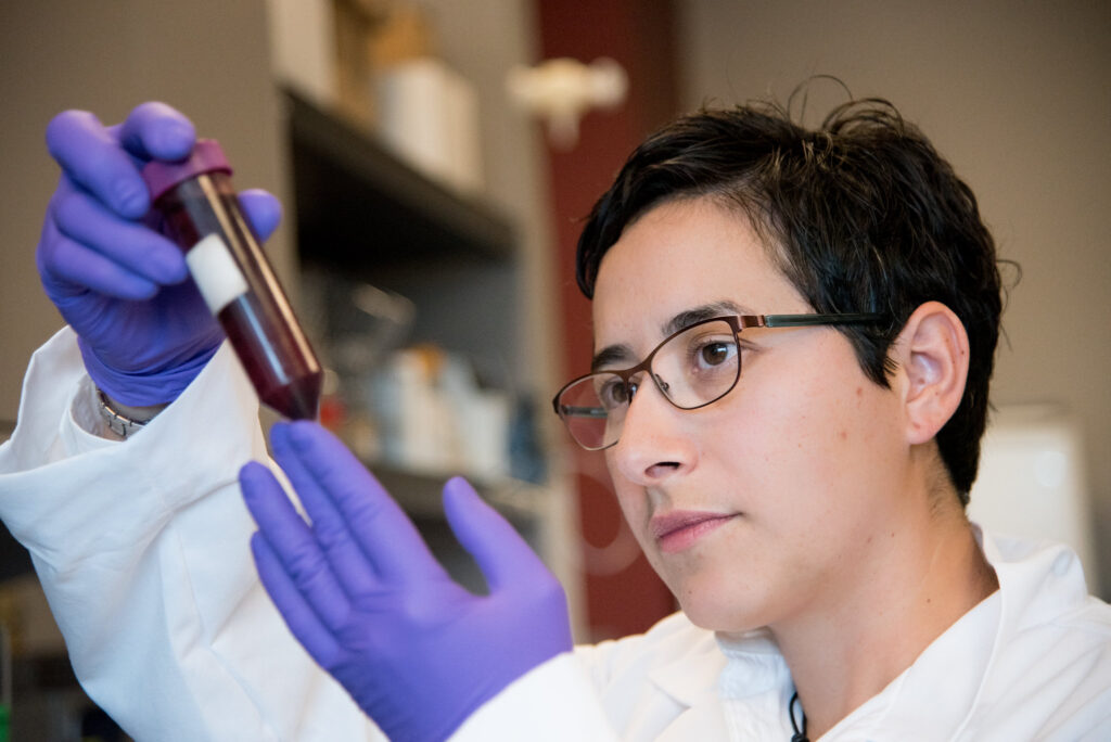 Student wearing a white lab coat and gloves examines a vial in her hands.