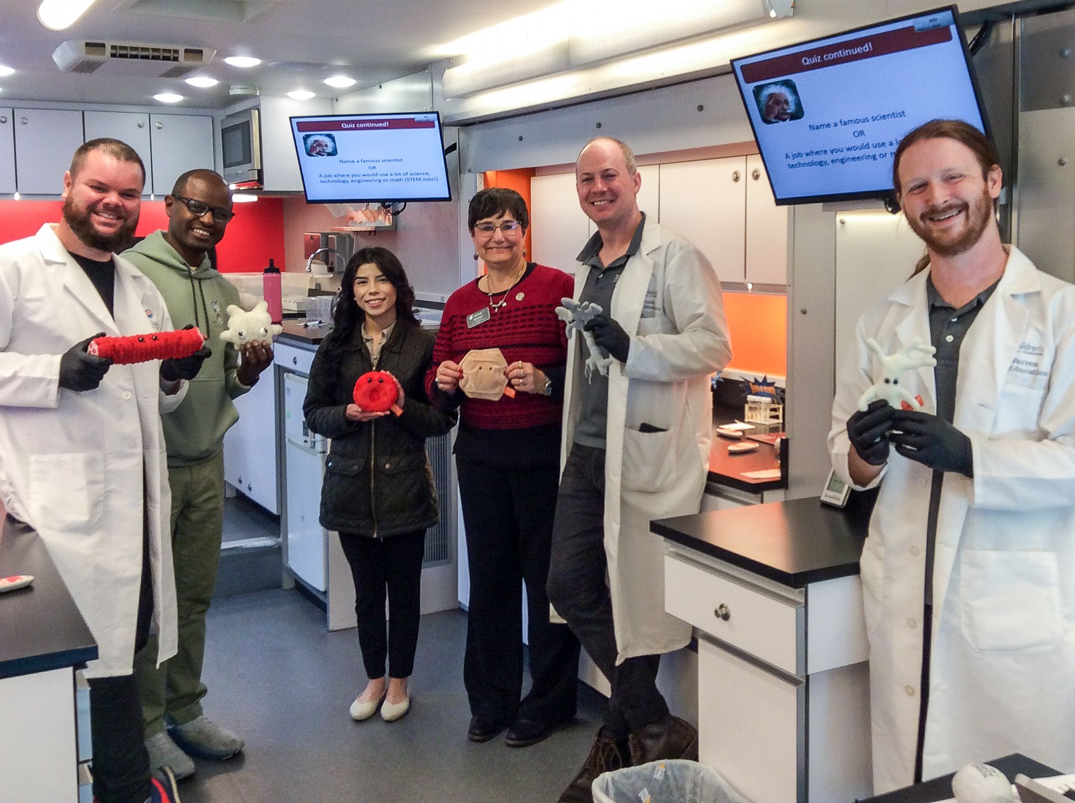 Lab leaders, Yonas, Yasmin and Noel standing inside the mobile lab holding plush toys in the shape of cells of the body.