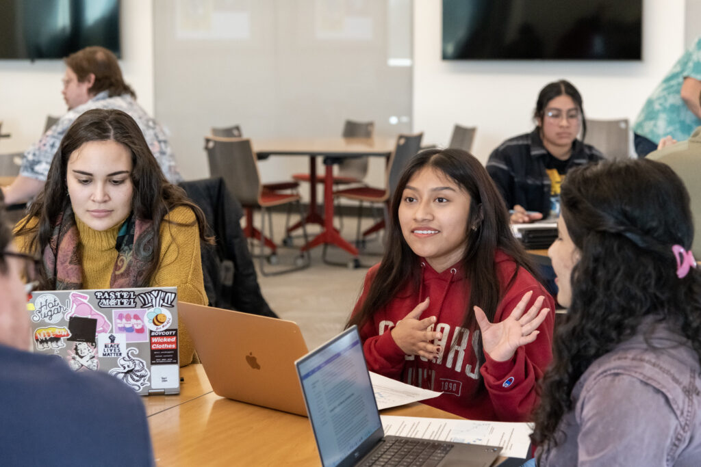 Students seated around a table with laptops talking to each other.
