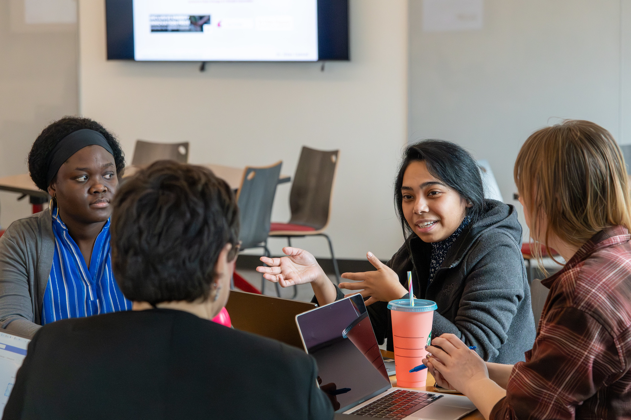 Students and mentors seated around a table with laptops talking to each other.