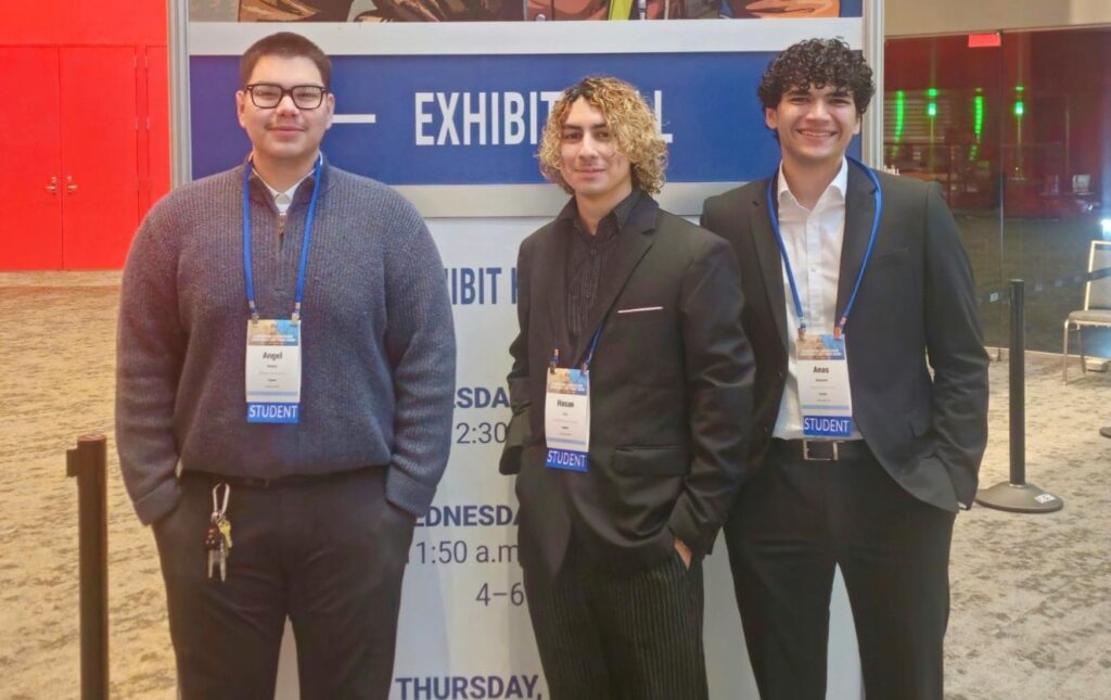 Three men in business casual attire stand in front of conference sign in hotel lobby.