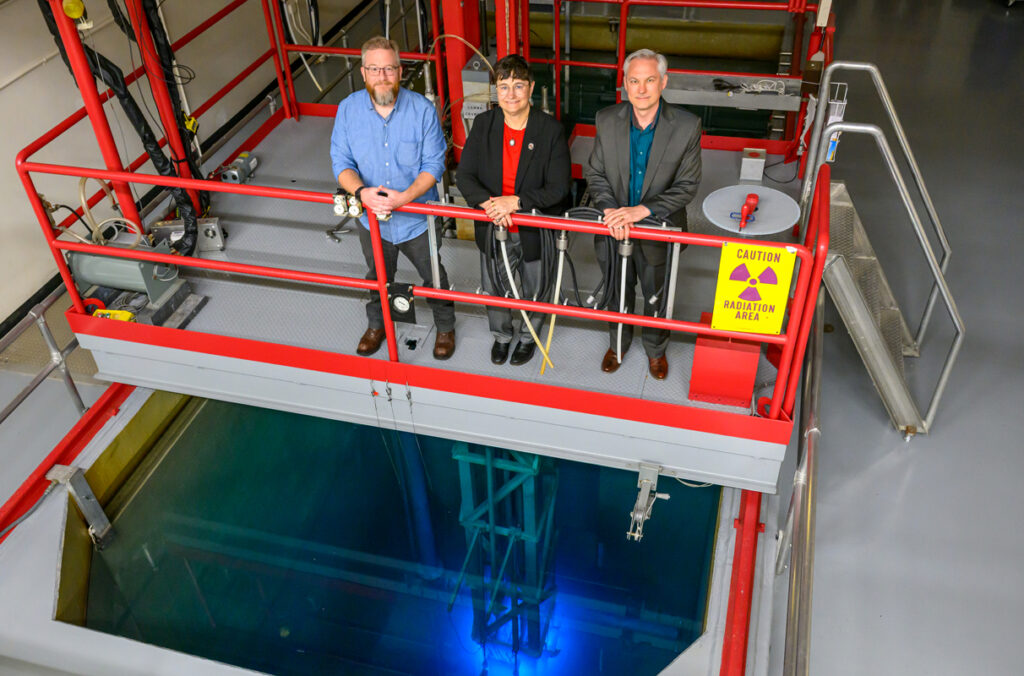 Three people stand on a platform overlooking an educational nuclear reactor.