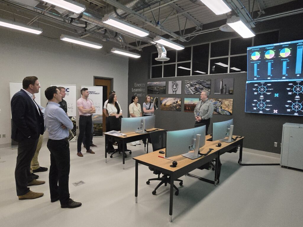Group of people stand in a room with computer three computer stations and large monitors.