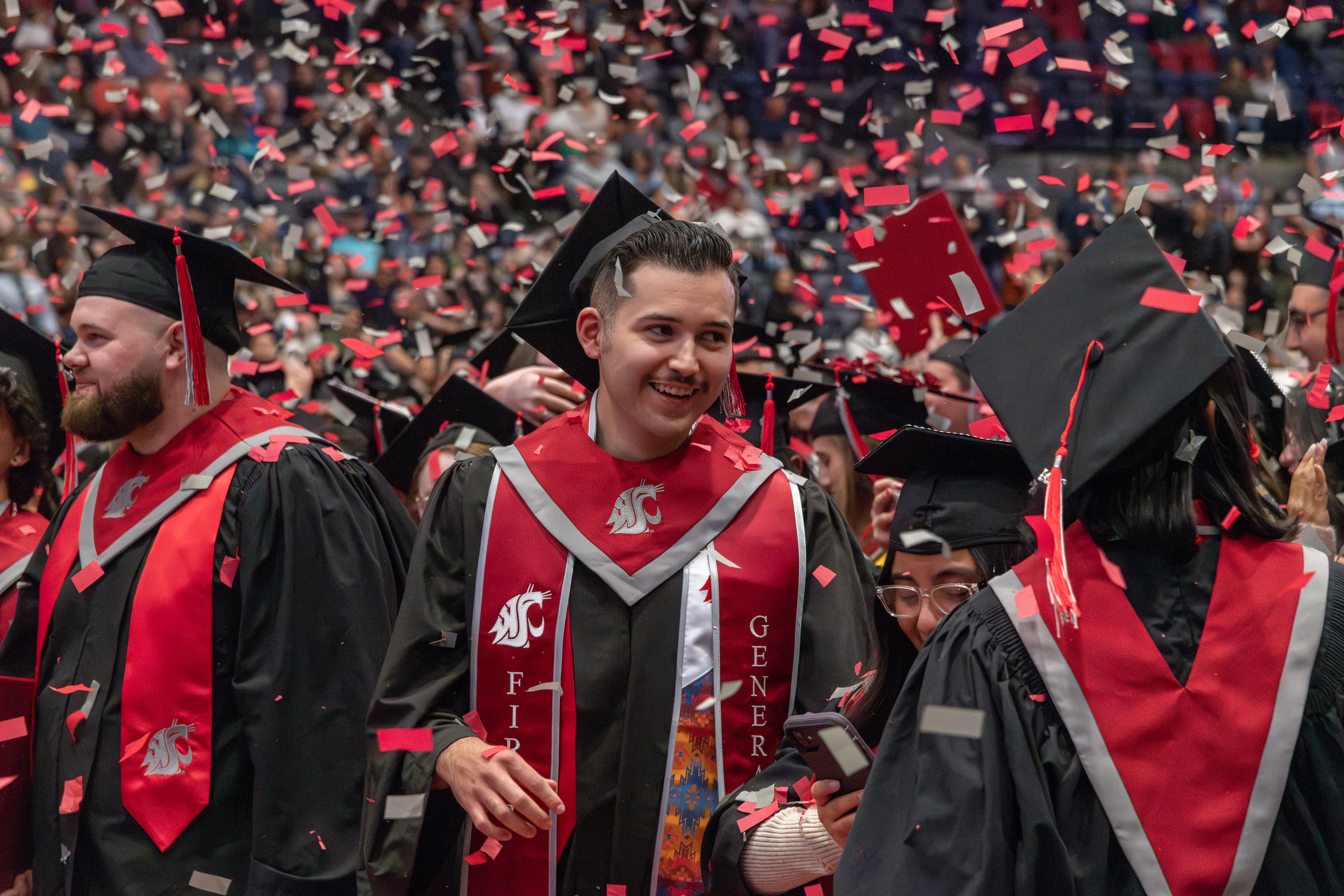 WSU Tri-Cities students wearing regalia and smiling as crimson and gray confetti falls on top of them.