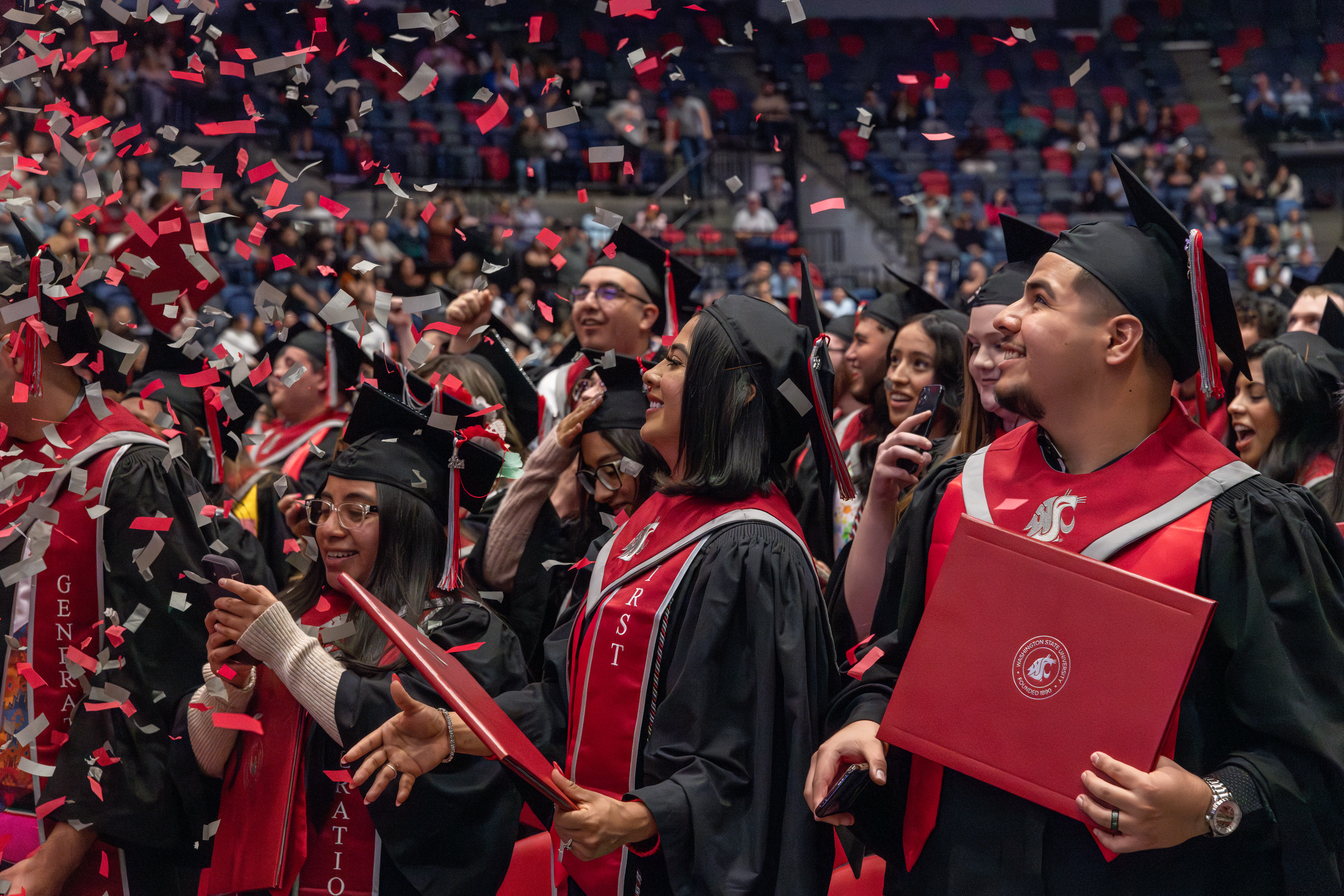A group of graduating WSU Tri-Cities students wearing regalia and holding diploma covers smiling as crimson and gray confetti falls in front of them.