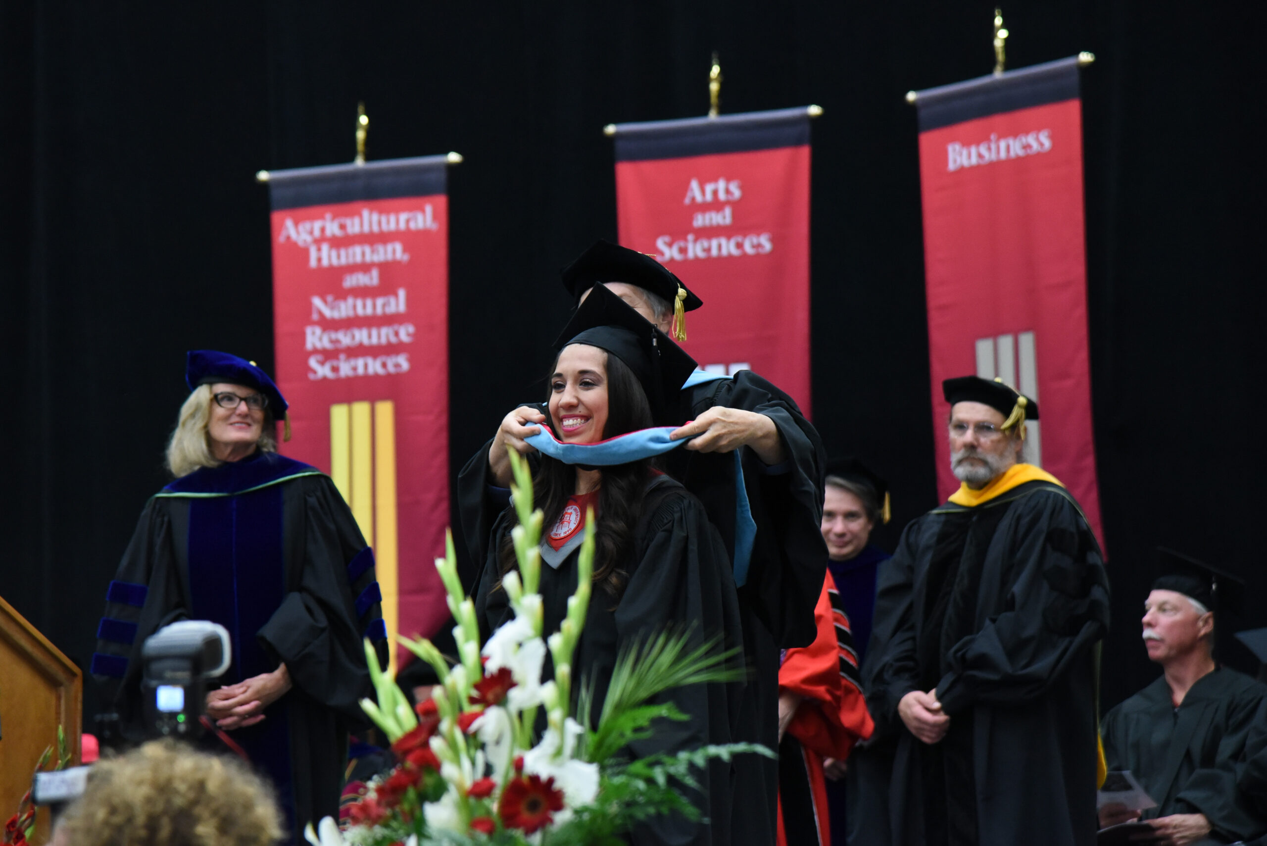 Graduating student wearing regalia on stage having a light blue hood being placed over her neck by a faculty member while others watch.