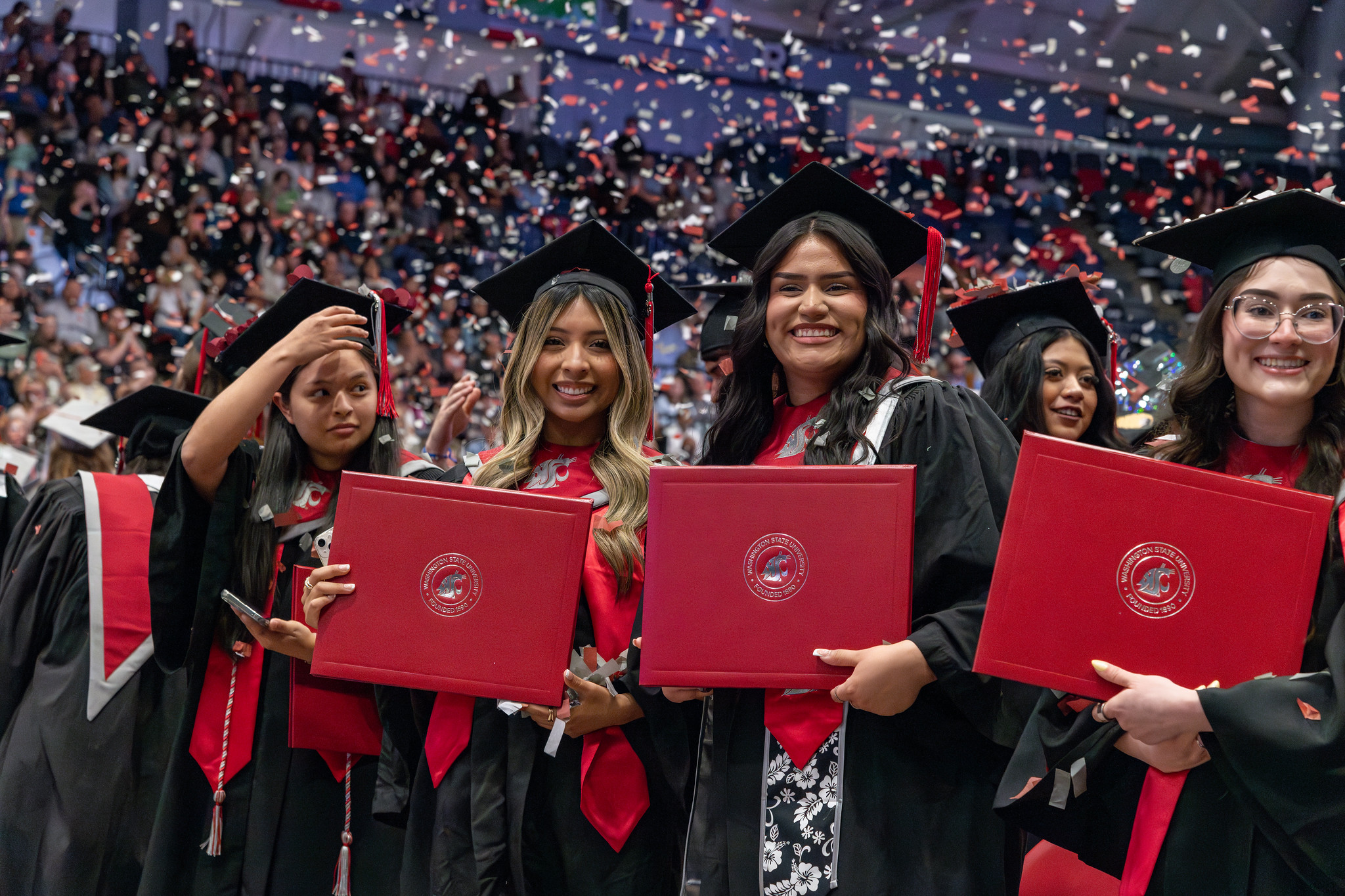 A group of graduates in caps and gowns holding WSU diploma covers in an arena as confetti falls from above.