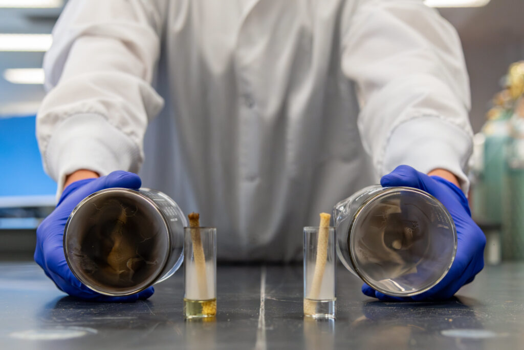 Person in lab coat holding two beakers next to each other to compare black smoke in one on the left and a clean beaker on the right.