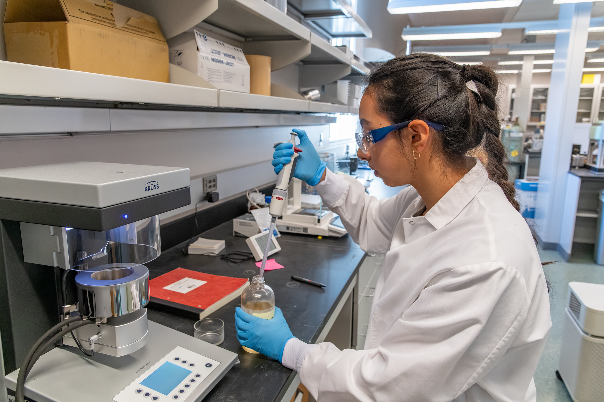 WSU Tri-Cities student in a white lab coat, gloves, and eye protection taking a sample of fuel from a bottle.
