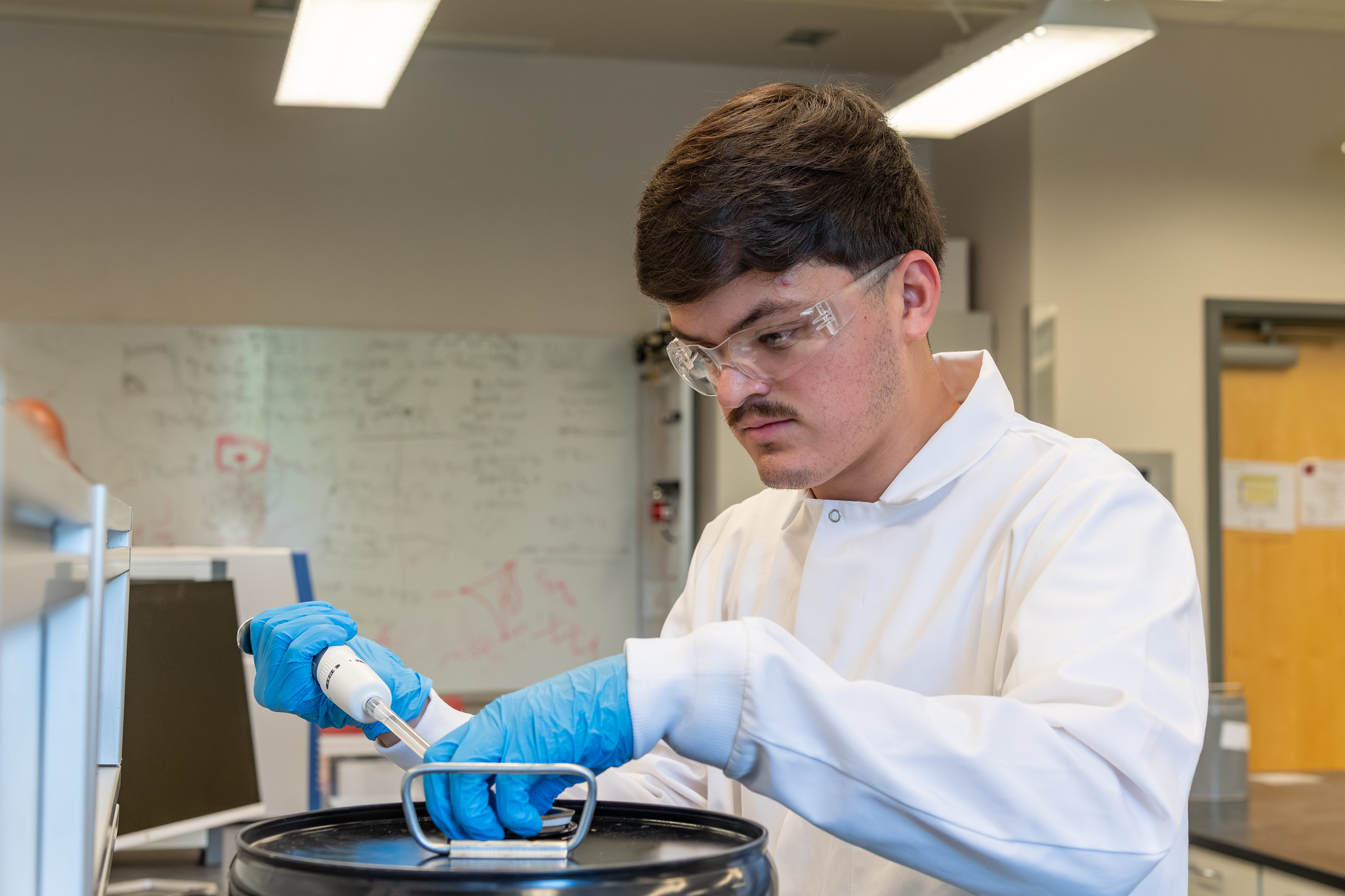 WSU Tri-Cities student in a white lab coat, gloves, and eye protection taking a sample of fuel from a black container.