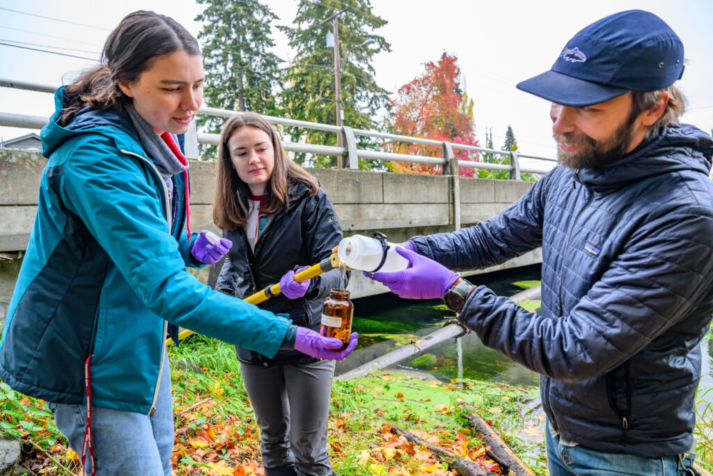 A person pouring liquid into a sample jar while another looks on.