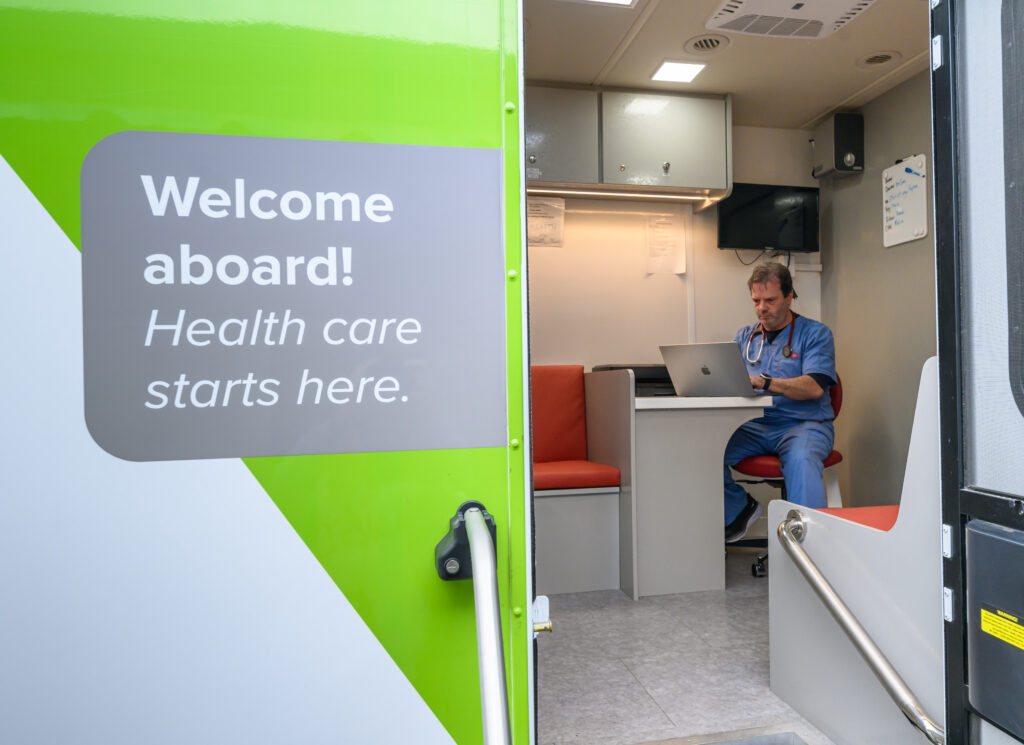 The open door of a mobile clinic truck, with a sign that reads "Welcome aboard! Health care starts here" and a doctor inside working on a laptop.