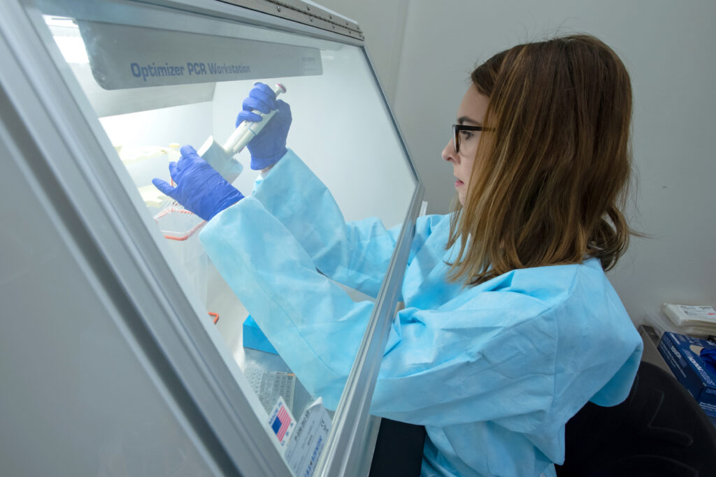 A microbiologist in blue protective equipment works at a biosafety cabinet.