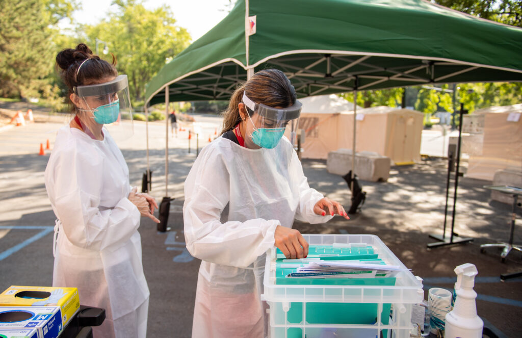 Two people in face masks and shields wearing full protective clothes working at an outdoor Covid-19 testing site in 2020.