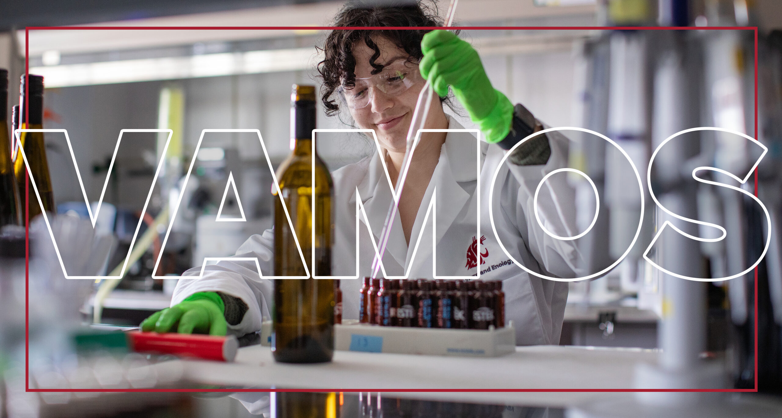 Texto grande “Vamos” superpuesto a un investigador sonriente con gafas de seguridad y una bata blanca de laboratorio, usando una herramienta para llenar botellas pequeñas con una botella de vino en primer plano.