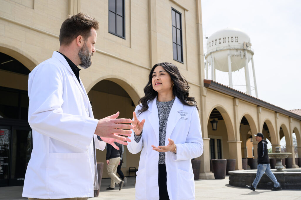 A pharmacy student wearing a white coat talking with another pharmacist, there is a tall water tower in the background with the word Toppenish.