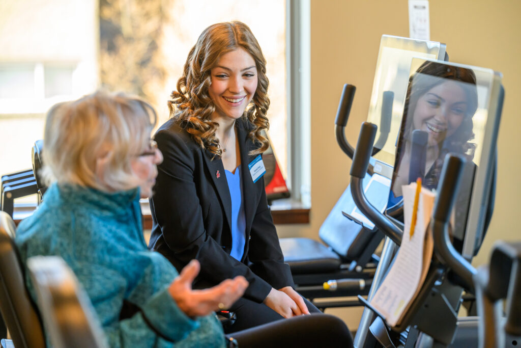 An older person sitting on an exercise machine and talking with a WSU graduate.