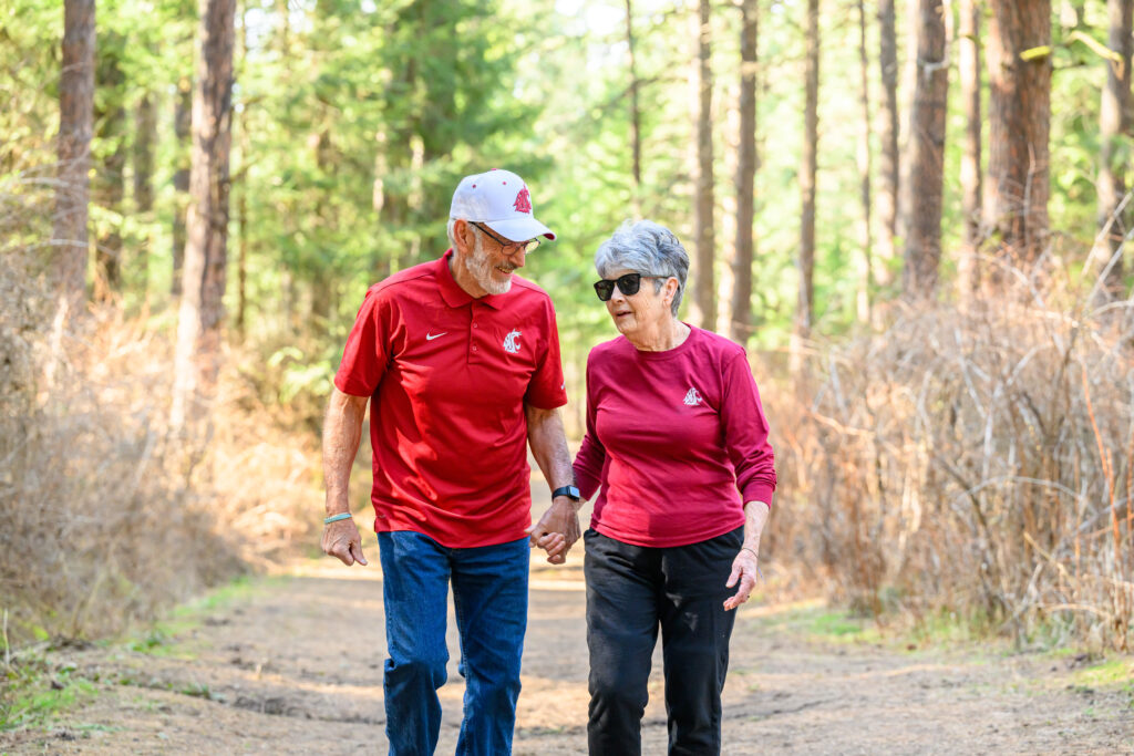 Two older adults, both wearing WSU Cougar branded shirts, holding hands on a walk through the woods.