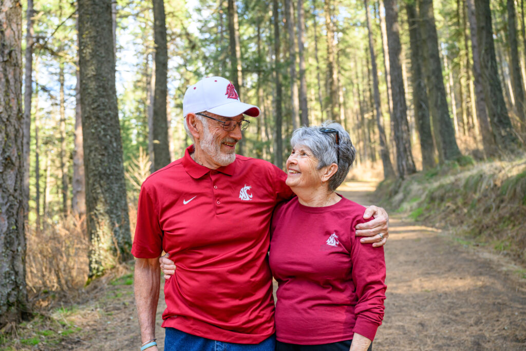 Two older adults, both wearing WSU Cougar branded shirts, smiling at each other as they walk together in the woods.